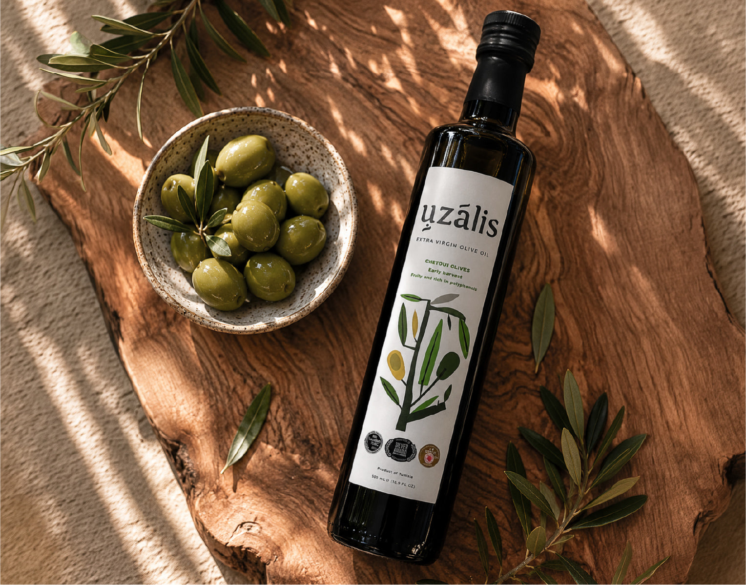 Uzalis Chetoui extra virgin olive oil bottle with fresh olives and Mediterranean ingredients on a rustic wooden board